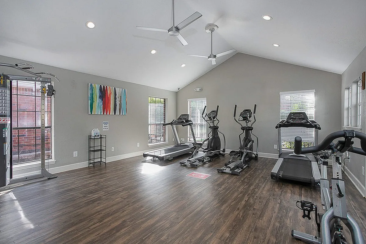 Modern fitness room at Autumn Ridge Apartments featuring sleek treadmills and ellipticals on polished wood floors with vibrant wall art and abundant natural light.