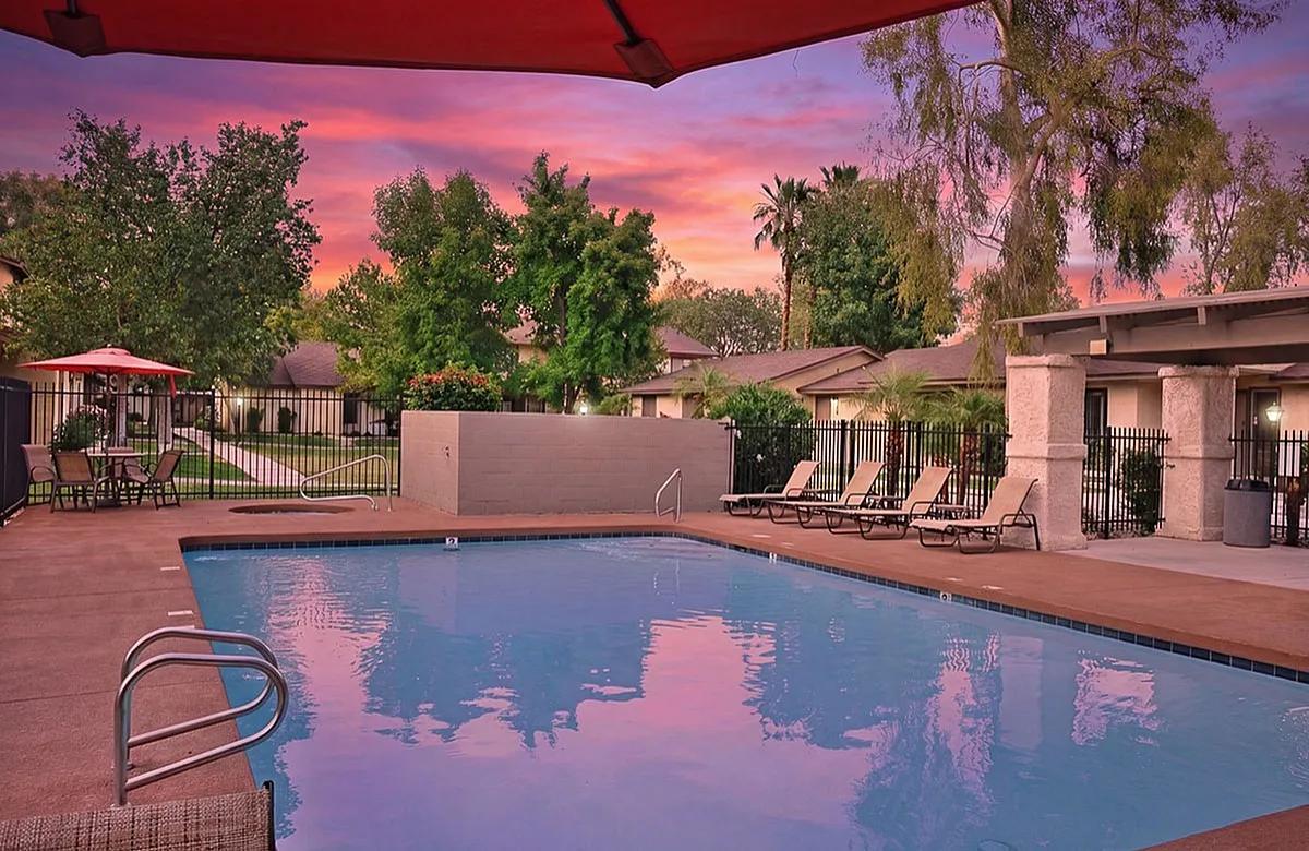Charming outdoor pool area at Camelot Apartments with comfortable lounge chairs, lush greenery, and a vibrant sunset sky at a beautiful apartment complex.