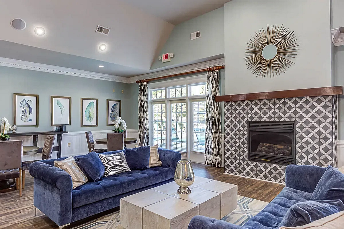 Elegant clubroom at The Stratford at Hillcrest Towne Center Apartments with plush blue sofas, geometric fireplace, modern decor, and large windows offering abundant natural light.