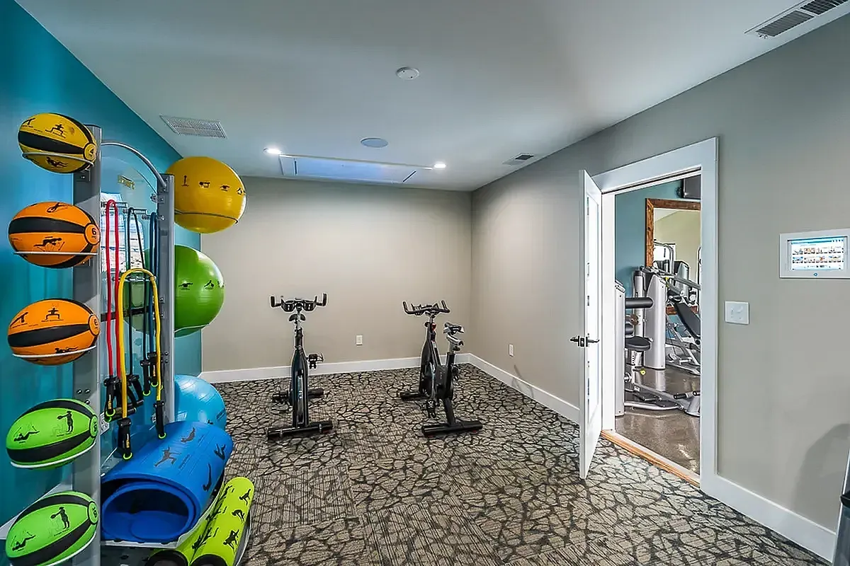 Inviting fitness room at The Colony at the Oaks Apartments with modern exercise bikes, vibrant medicine balls, and an alluring beach artwork for a refreshing workout ambiance.