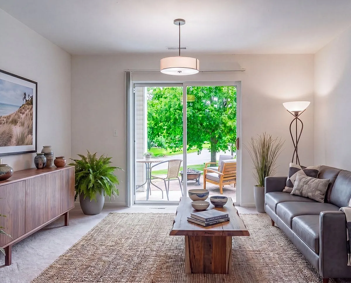 Inviting living room with elegant decor, featuring a cozy gray sofa, chic wooden coffee table, and vibrant greenery seen through glass doors leading to a sunny patio Clearview Apartments in Holland, MI