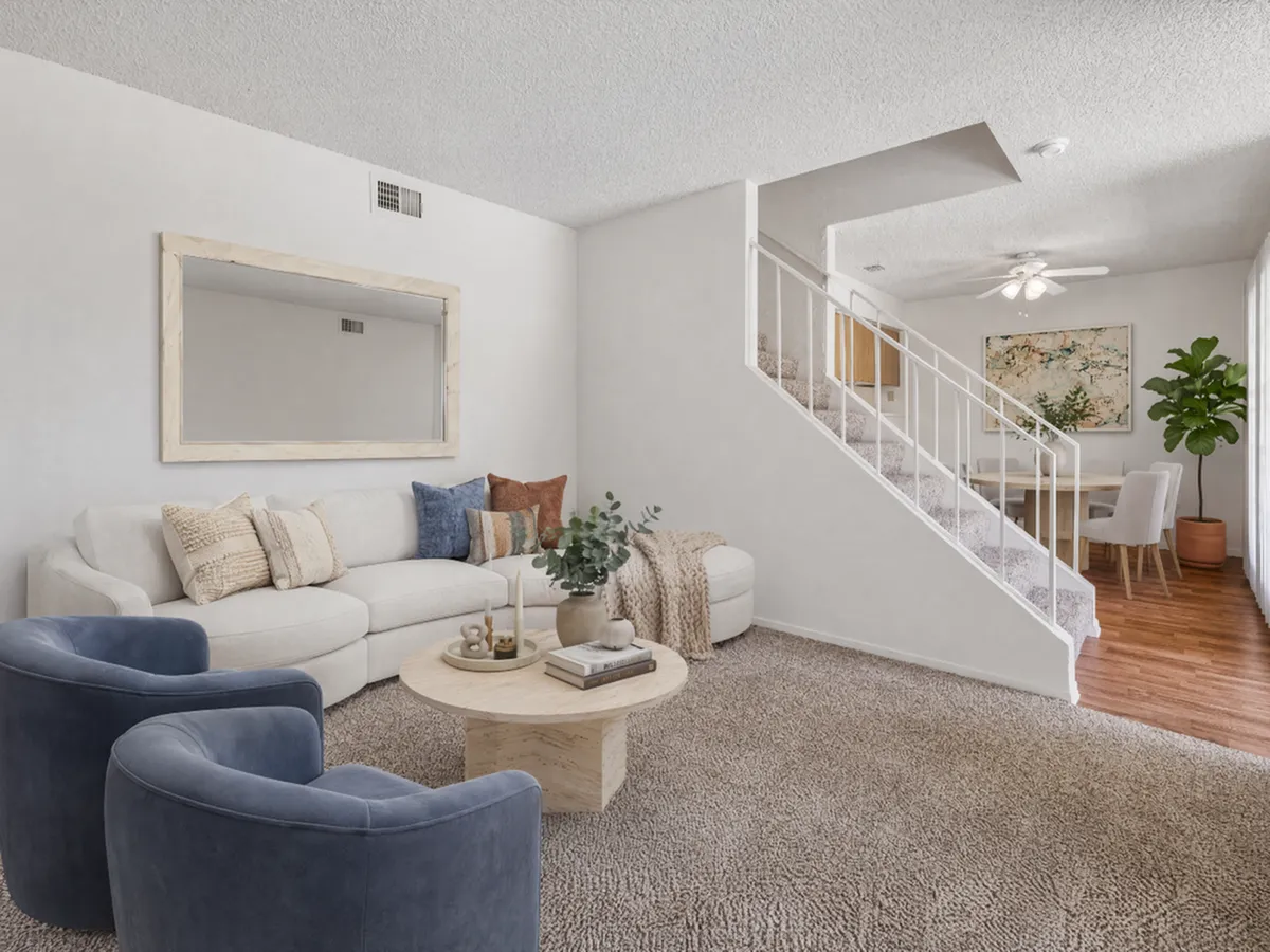 Inviting living room with elegant beige sofa, plush blue chairs, and stylish decor leading to a charming dining area with natural light and a modern staircase at Camelot Apartments