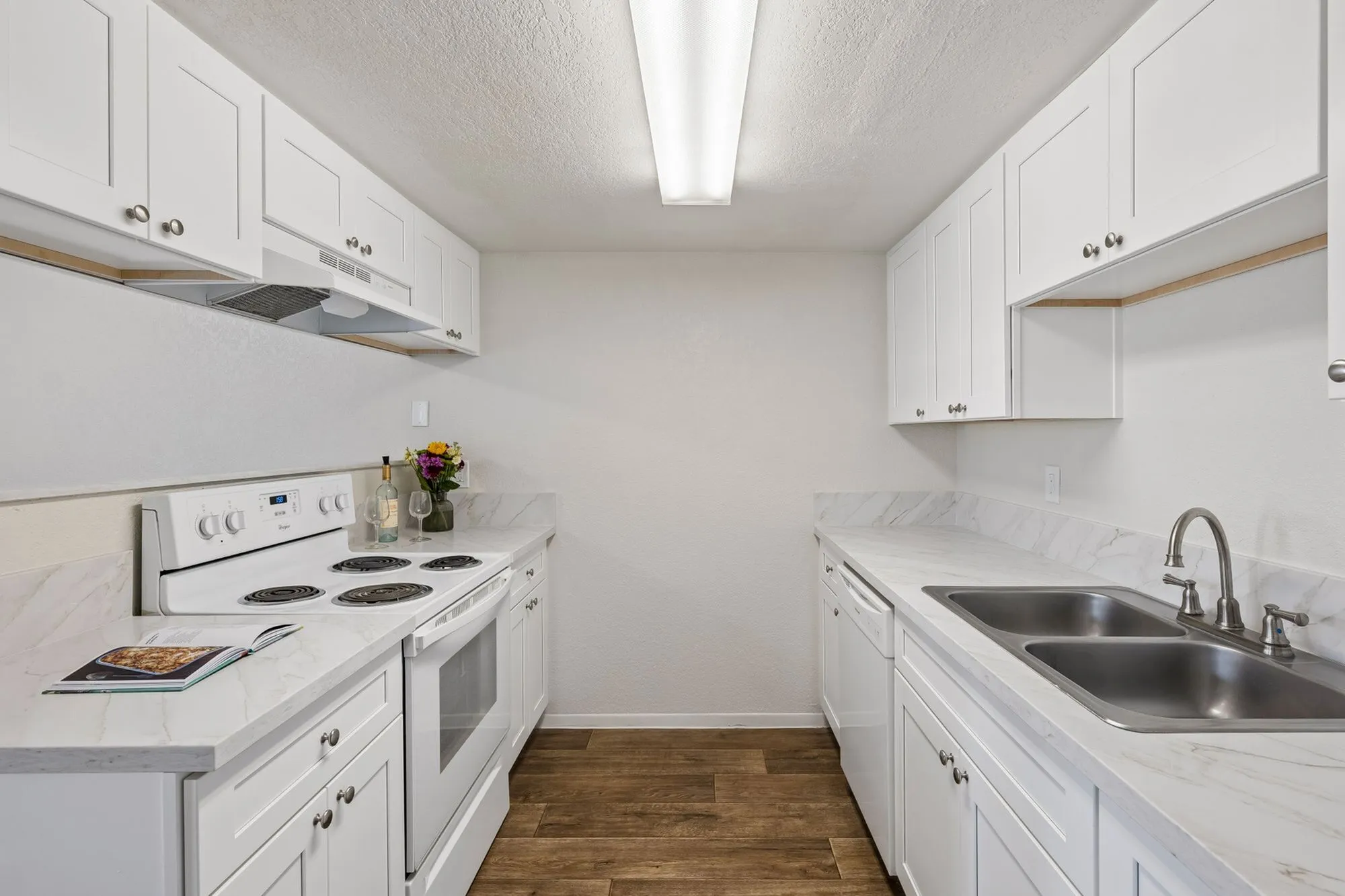 Modern and inviting kitchen featuring sleek white cabinetry, marbled countertops, and stainless steel sink with elegant wood flooring at Camelot Apartments in Yuma, AZ.