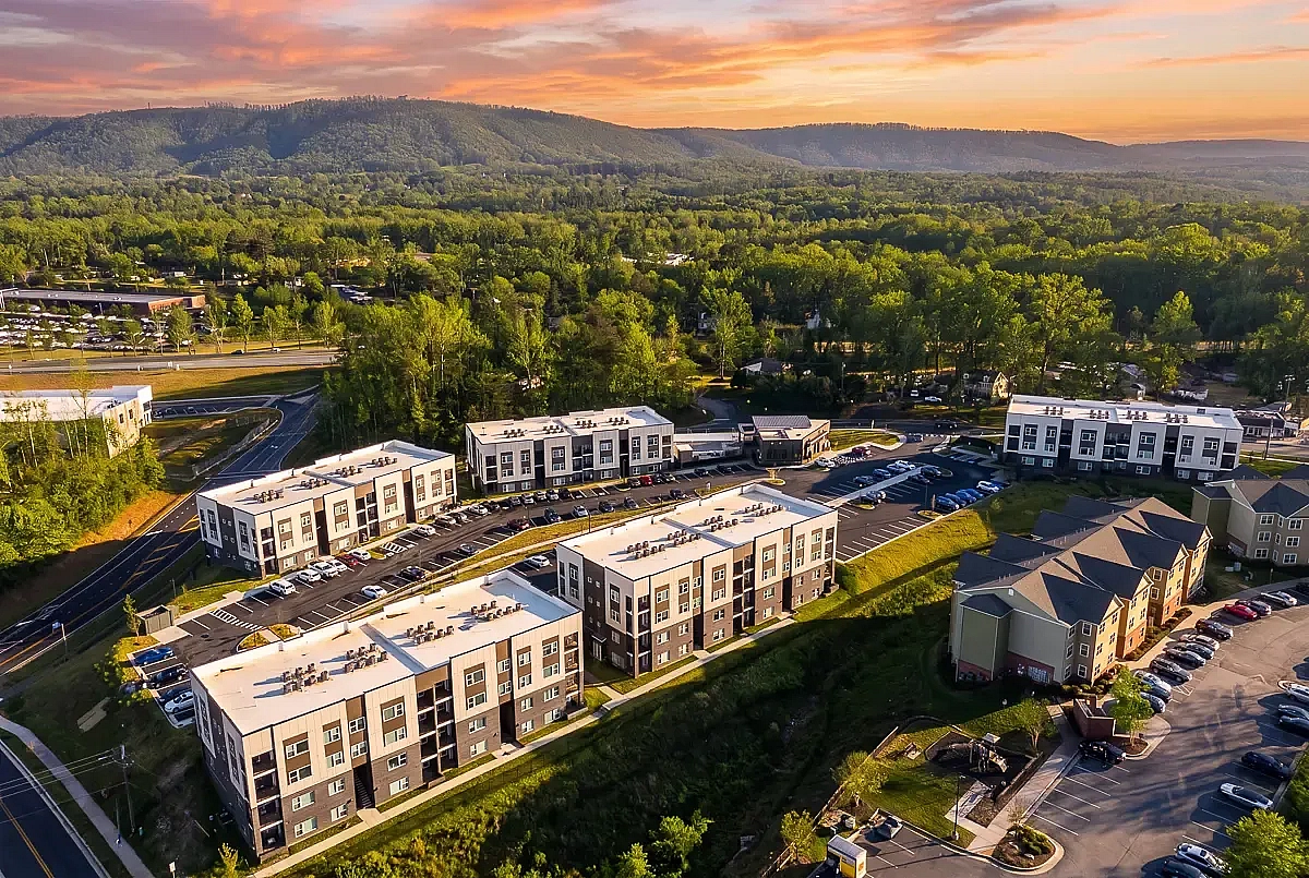 Aerial view of modern apartment complex in Charlottesville, VA, nestled amidst lush greenery with a stunning mountain backdrop and vibrant sunset sky.