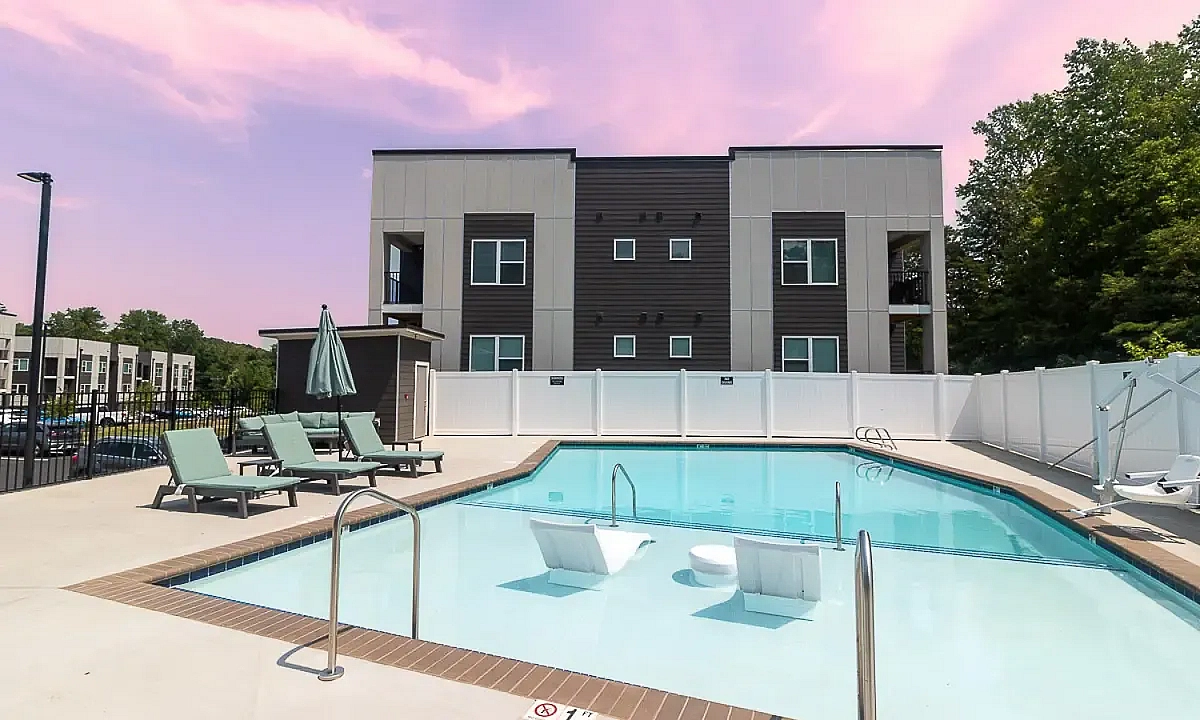 Modern apartment complex in Charlottesville with a luxurious outdoor pool, surrounded by lounge chairs and green trees, under a vibrant pink sky at 5 Row Apartments.