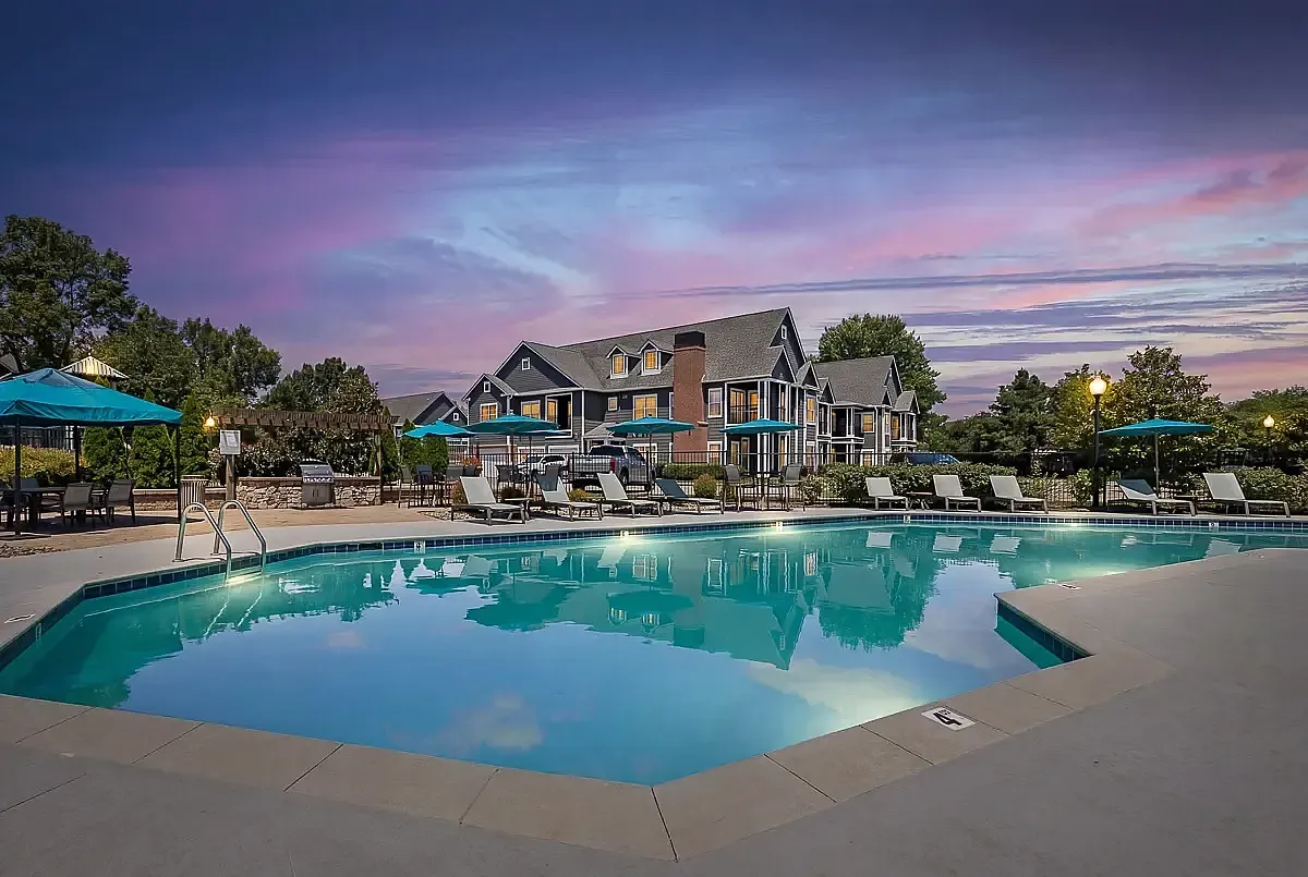 Luxurious apartment complex at sunset with a sparkling pool, modern loungers, and ambient lighting, set against a stunning sky.
