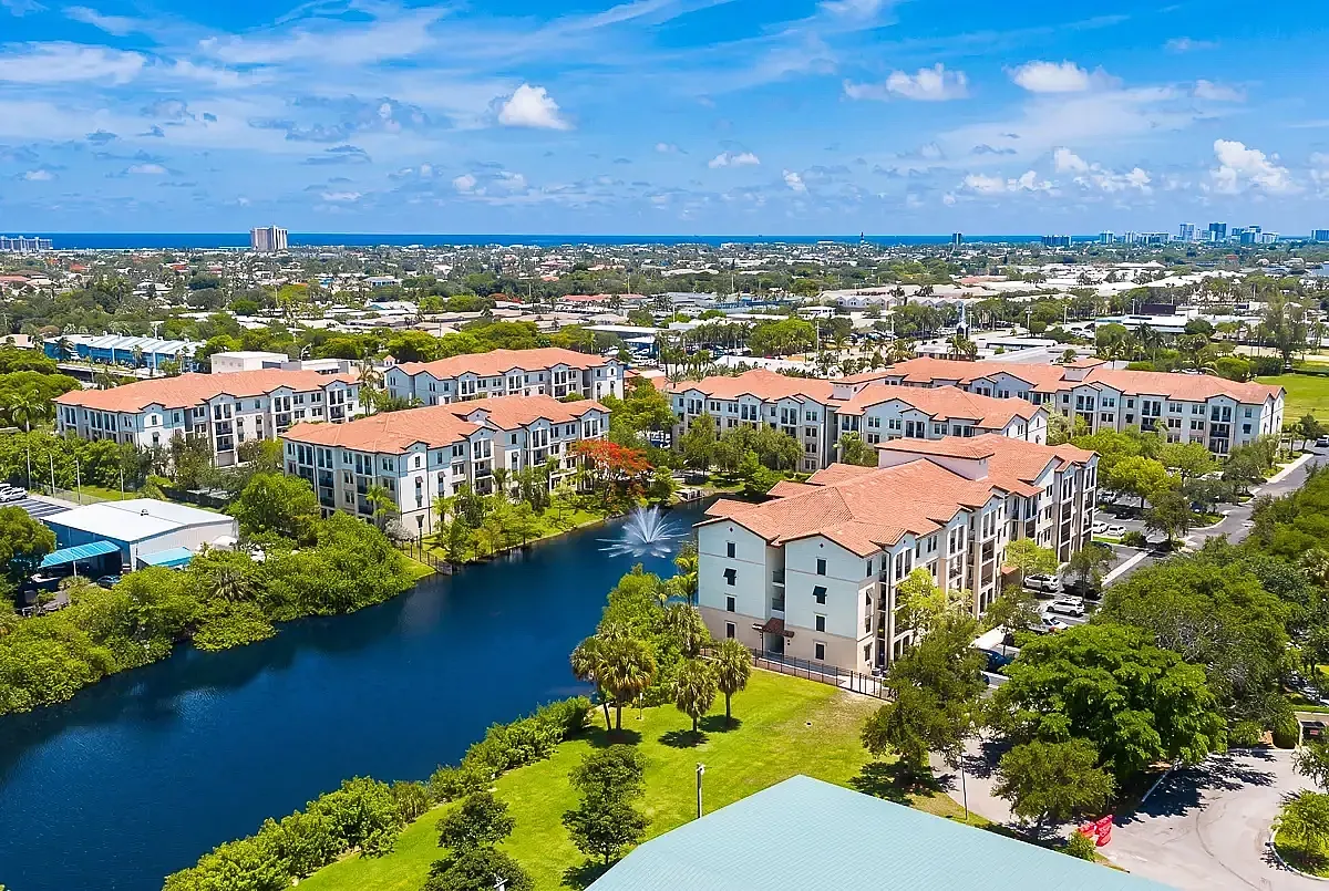Elegant aerial view of modern apartment complex with Mediterranean-style roofs, lush green surroundings, and a serene lake with a sparkling fountain, set against a vibrant cityscape and clear blue sky.