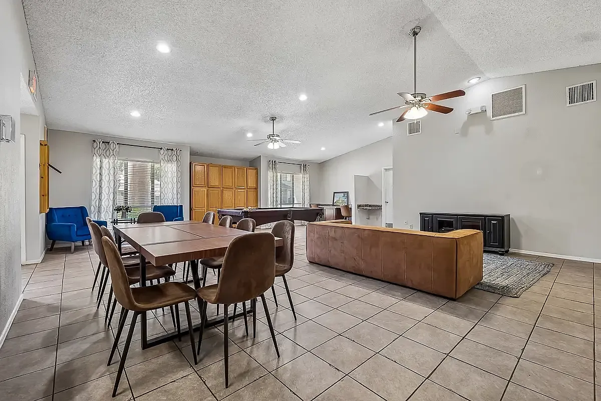 Spacious open-concept living area with modern dining set, cozy brown couch, large wall-mounted TV, and bright natural lighting at Camelot Apartments in Yuma, AZ.