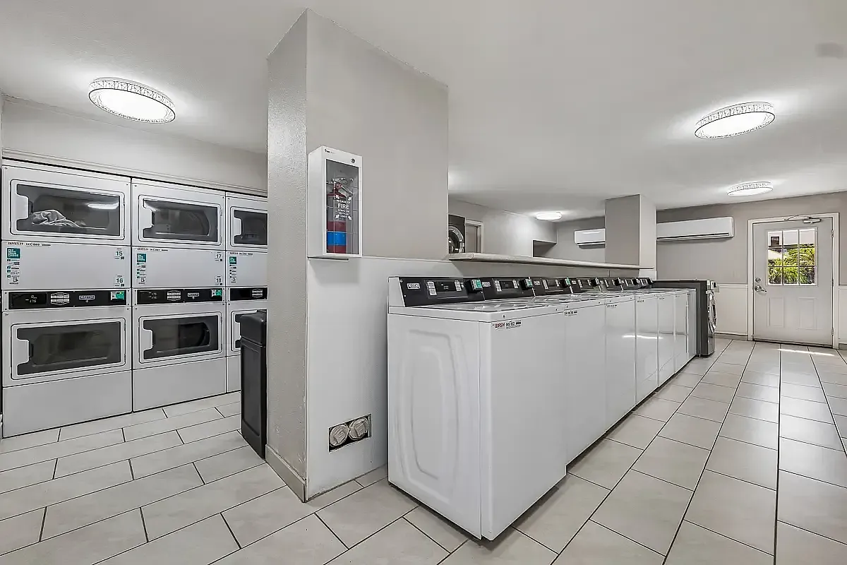 Bright and modern communal laundry room featuring multiple washers and dryers, sleek tile flooring, and ample lighting for convenience  at Camelot Apartments in Yuma, AZ.