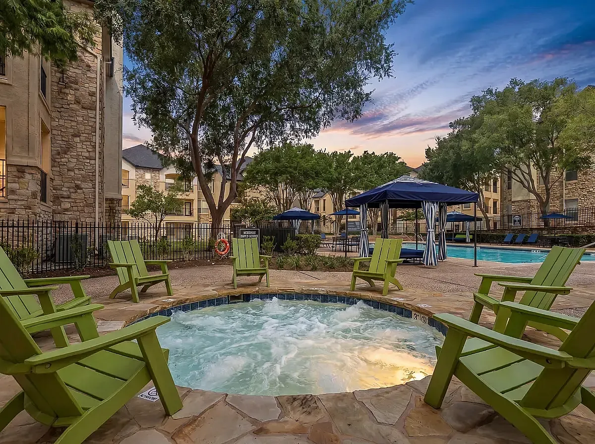 Inviting outdoor oasis at Stonebriar of Frisco Apartments featuring a luxurious hot tub surrounded by charming green chairs and elegant cabanas against a picturesque sunset backdrop.