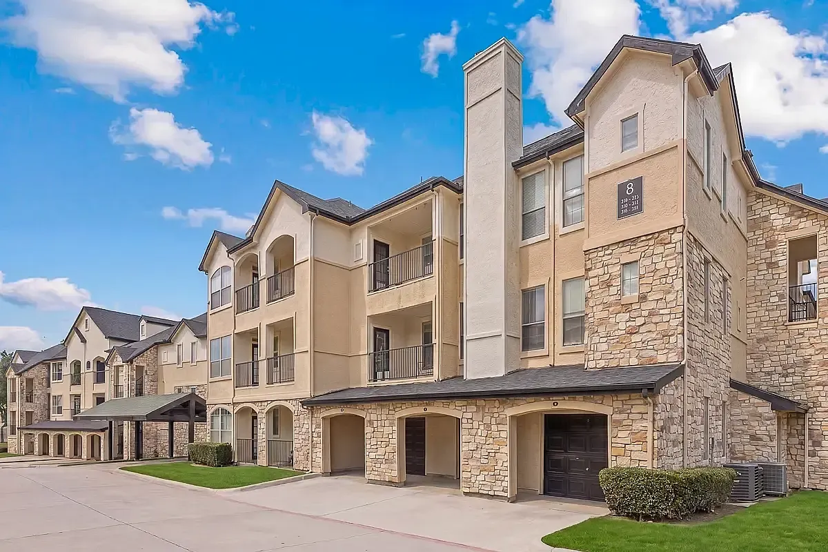 Charming three-story apartment building, Stonebriar of Frisco Apartments, with elegant stone facade and covered balconies, situated in a serene neighborhood under a clear blue sky.