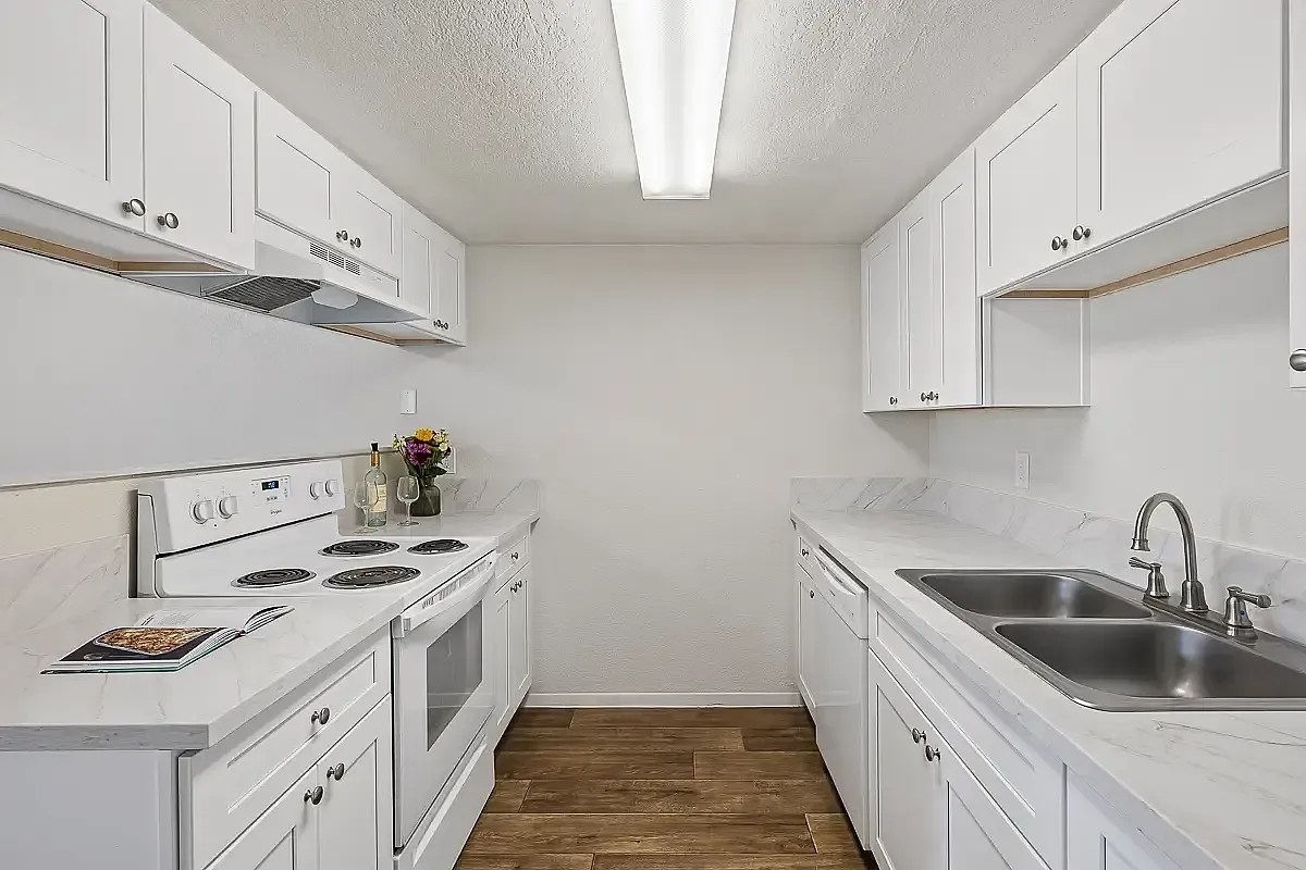 Modern and inviting kitchen featuring sleek white cabinetry, marbled countertops, and stainless steel sink with elegant wood flooring at Camelot Apartments in Yuma, AZ.