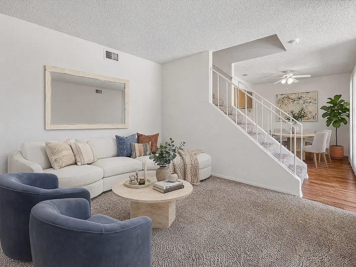 Inviting living room with elegant beige sofa, plush blue chairs, and stylish decor leading to a charming dining area with natural light and a modern staircase at Camelot Apartments