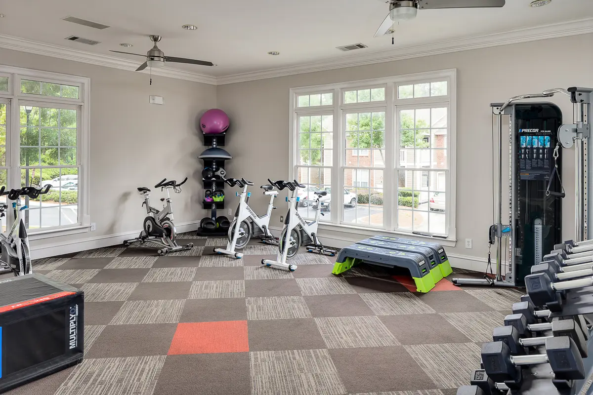 Contemporary gym at Retreat at Peachtree City Apartments with exercise bikes, weights, and modern equipment set in a bright room with large windows.