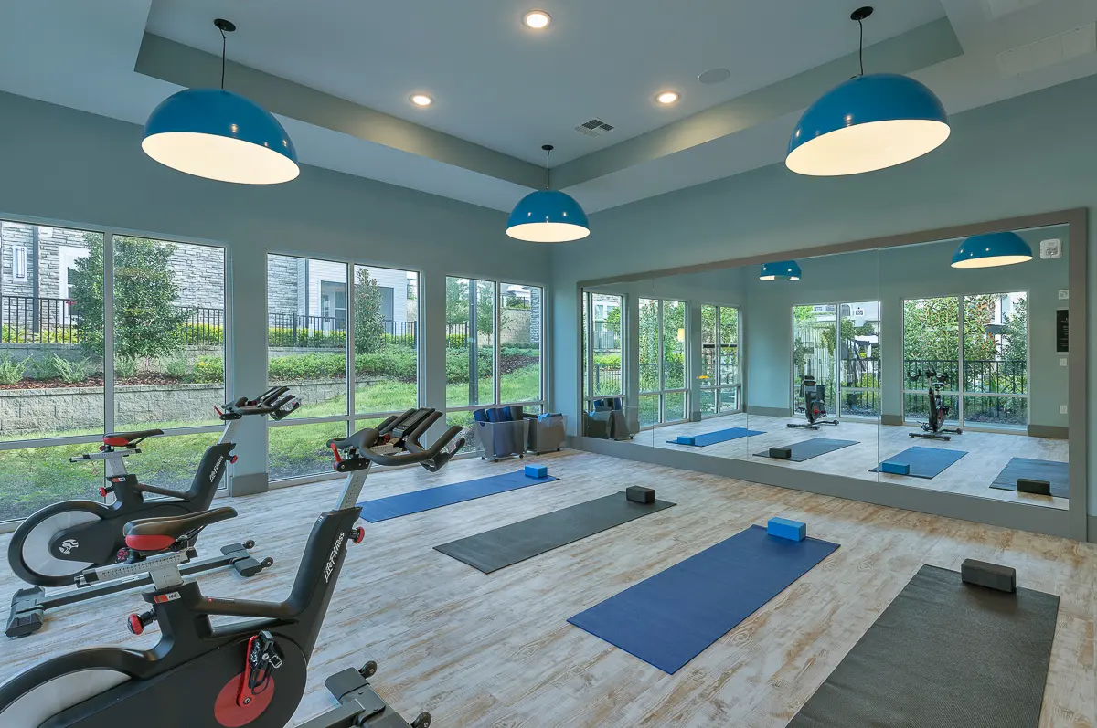 Modern gym facility at Cottonwood Clermont Apartments with large windows and cityscape views, featuring exercise bikes, yoga mats, and vibrant blue lighting.