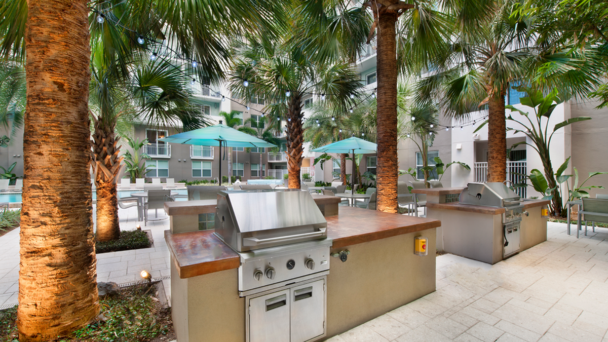 Luxurious outdoor courtyard with palm trees, modern grill stations, and chic seating areas under teal umbrellas, perfect for social gatherings in Downtown St Pete