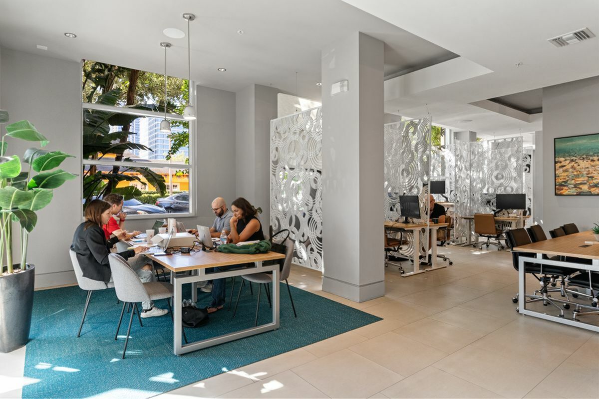 Modern open-concept office space with stylish decor, featuring collaborative workstations and abundant natural light from large windows at Apt CoWork in downtown st petersburg at Cottonwood Bayview Apartments