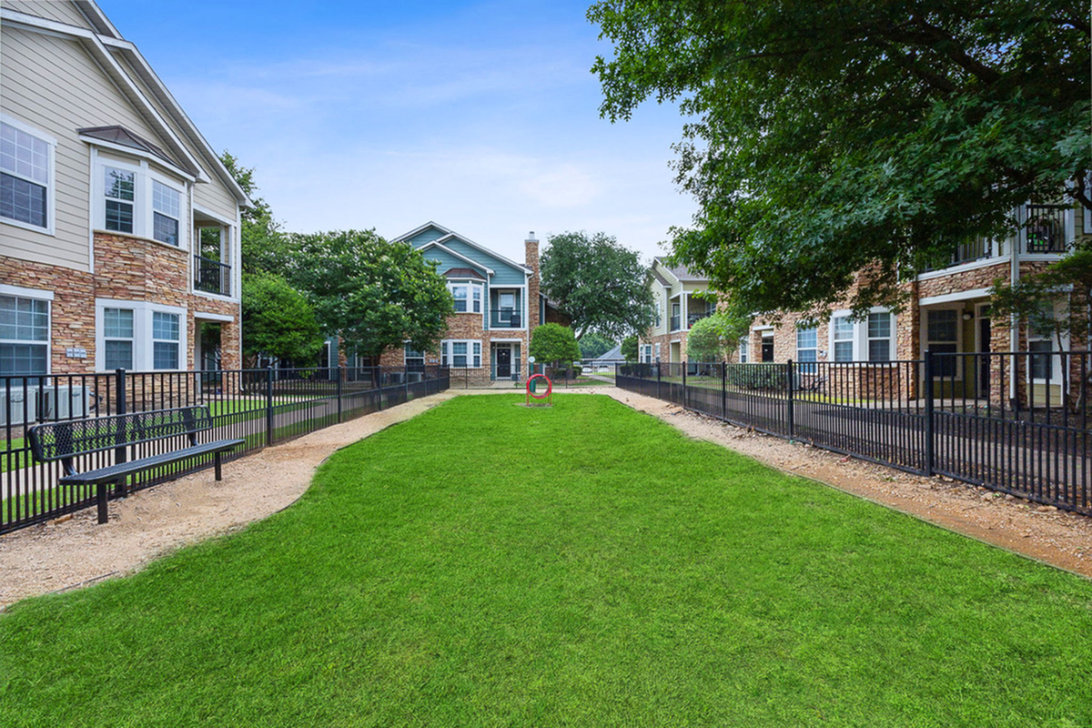 Charming dog park at Cottonwood Ridgeview Apartments in Plano with well-maintained green lawn, surrounded by picturesque buildings and lush trees, offering a peaceful and inviting community atmosphere.