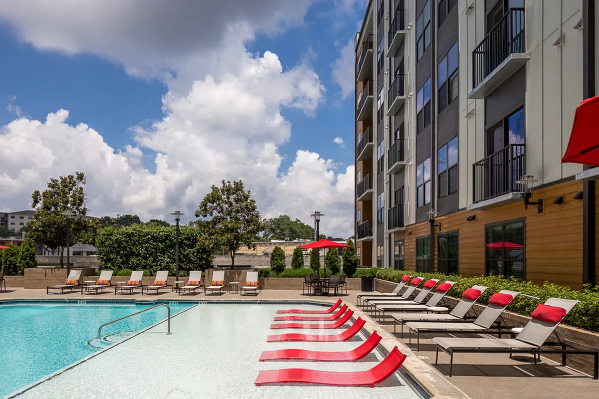 Inviting apartment pool at Cottonwood Westside with modern red lounge chairs, surrounded by lush greenery and stylish apartment building under a bright, blue sky.