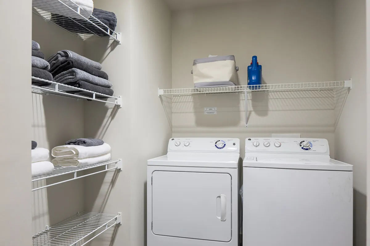 Modern laundry room at Cottonwood Westside Apartments equipped with a washer and dryer alongside organized shelving stocked with neatly folded towels, offering both convenience and style.