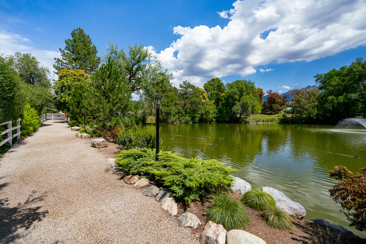 Scenic lakeside path at Fox Point in Old Farm Apartments with lush greenery, tranquil water, and a picturesque fountain beneath a vibrant blue sky.
