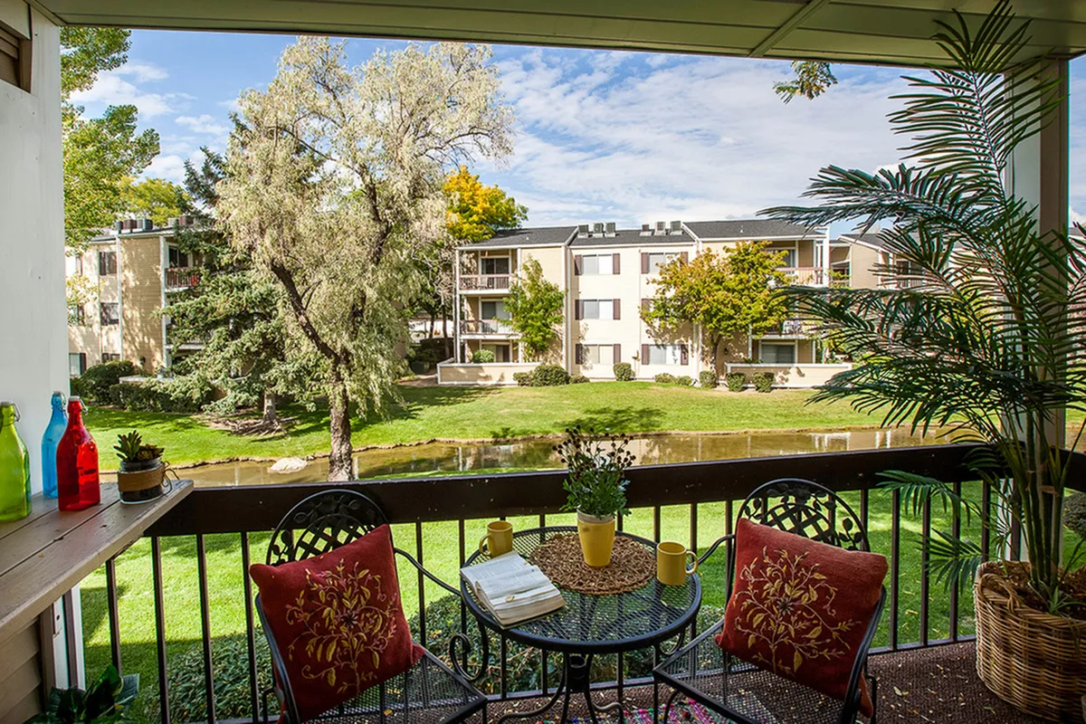 Charming balcony view at Fox Point in Old Farm Apartments featuring vibrant greenery, a serene pond, and cozy outdoor seating with decorative cushions in a stylish apartment complex.