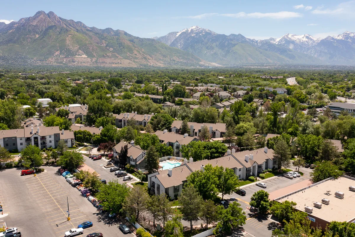 Scenic aerial view of a vibrant apartment complex surrounded by lush greenery and majestic mountains, featuring a sparkling shared pool and ample parking.