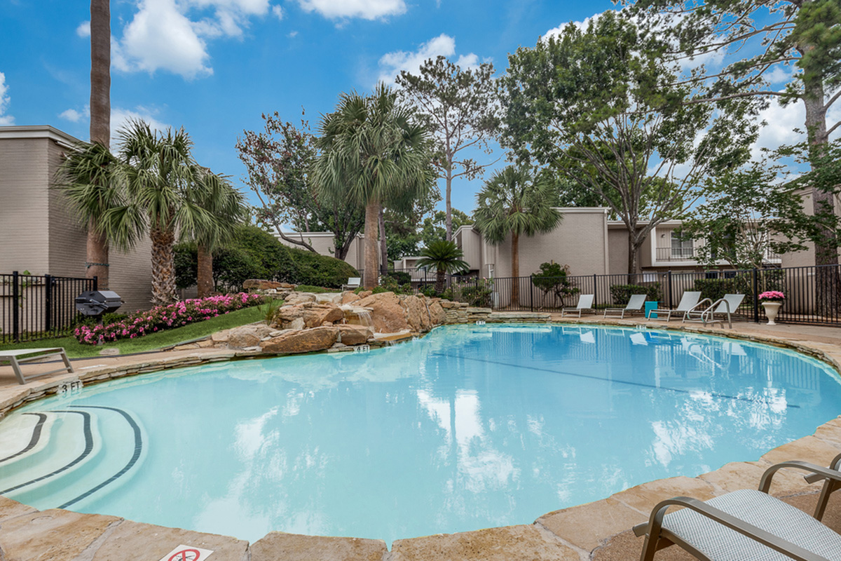 Tranquil apartment pool area surrounded by lush greenery and elegant palm trees, featuring comfortable lounge chairs and a serene atmosphere.
