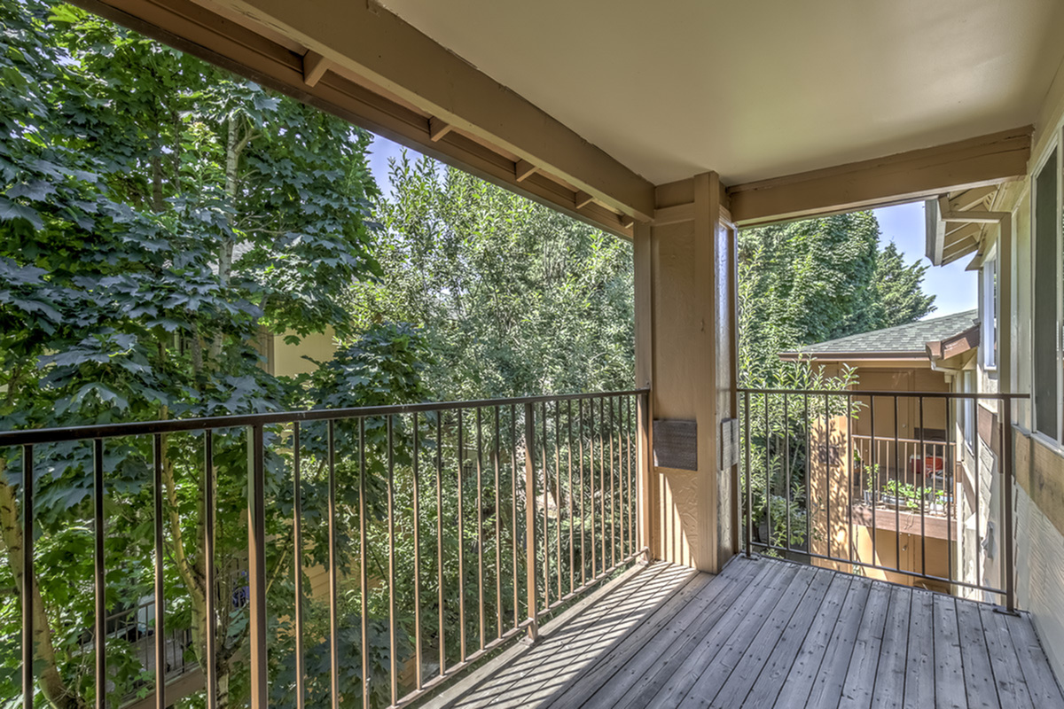 Charming apartment balcony with wooden flooring, offering a tranquil view of lush green trees and neighboring buildings.