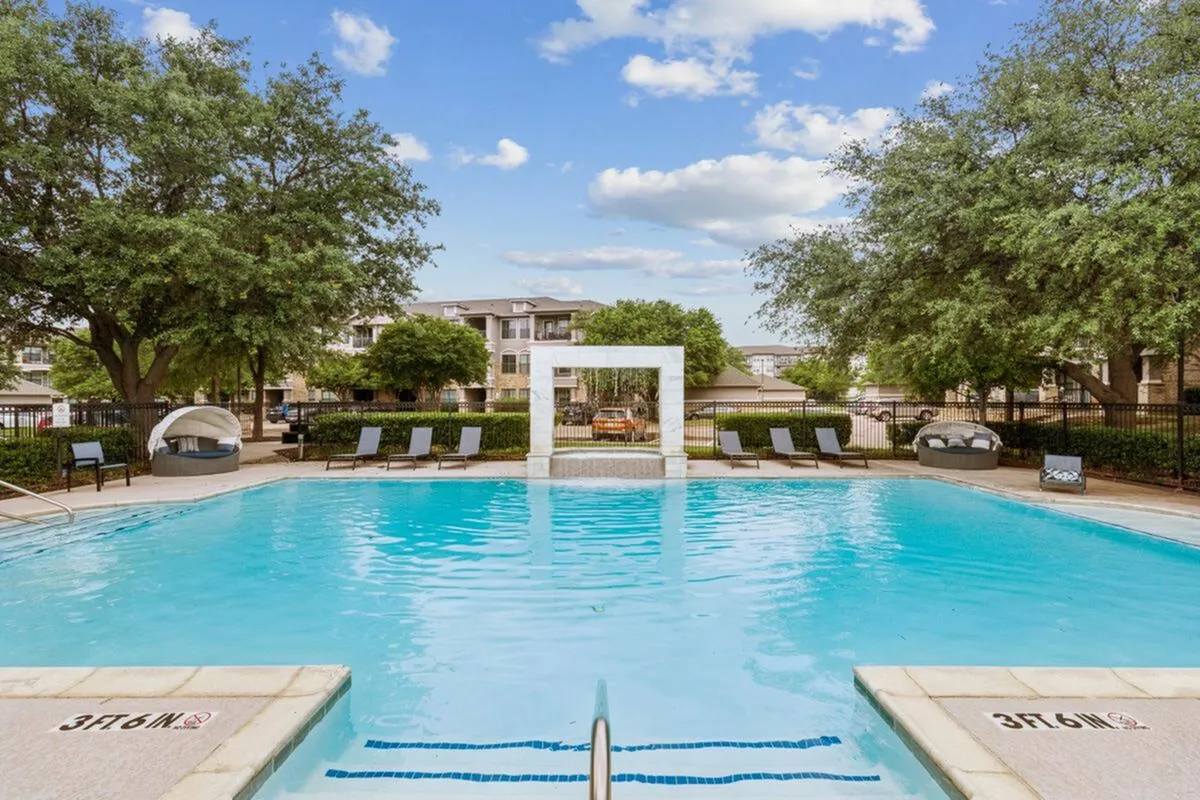Inviting outdoor pool area surrounded by lush greenery, elegant lounge chairs, and cozy cabana seating, under a bright blue sky.