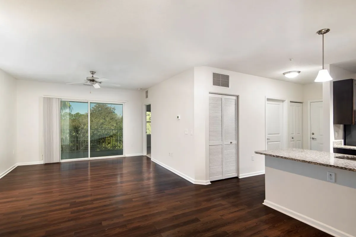 Spacious open-plan living room with dark hardwood floors, a ceiling fan, and a large sliding glass door leading to a scenic balcony. Modern kitchen with granite countertops adds elegance.