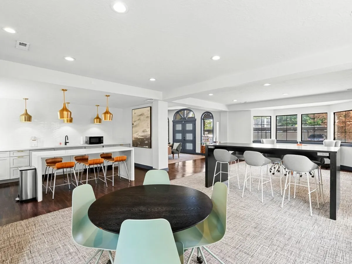 Bright, open-concept living area featuring a modern kitchen with gold pendant lighting, sleek white countertops, and stylish bar stools. Spacious dining and lounge spaces with large windows offering natural light at Cottonwood Apartments in SLC