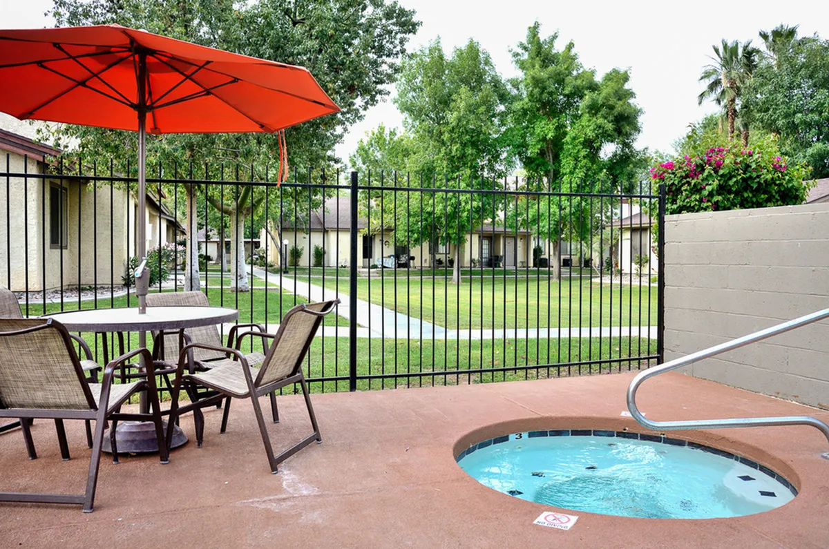 Charming outdoor patio with cozy seating, vibrant red umbrella, and inviting hot tub overlooking lush gardens and well-maintained walkways.