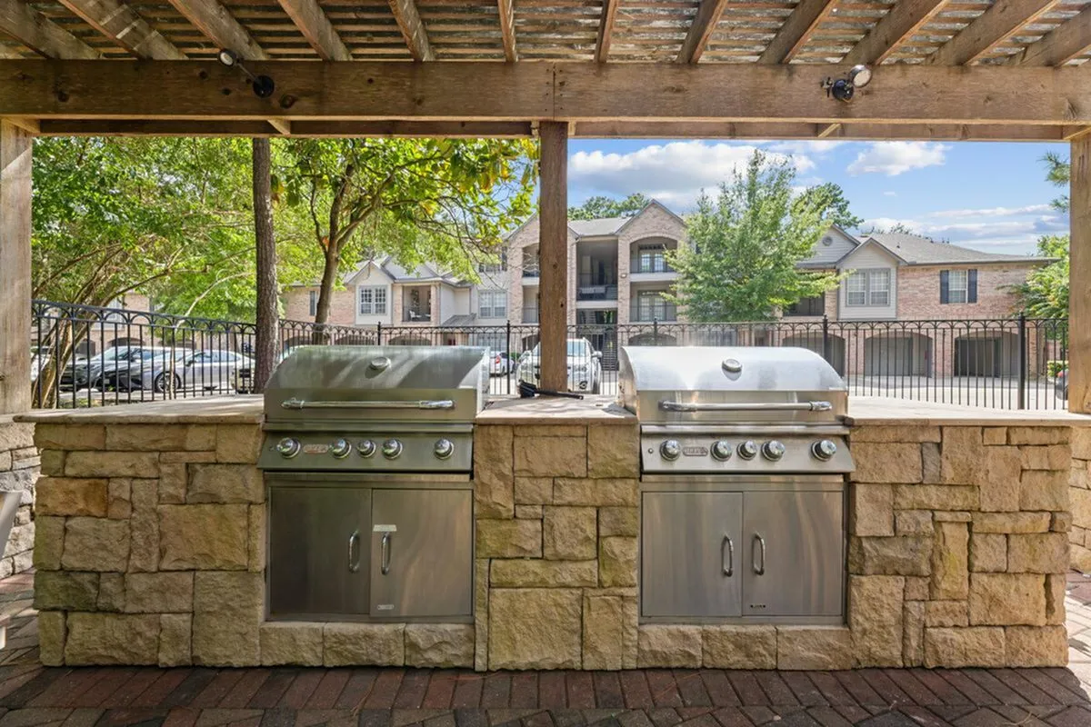 Charming outdoor barbecue area with two stainless steel grills under a rustic pergola, surrounded by lush greenery and elegant apartment building at Raveneaux Apartments in Houst Texas