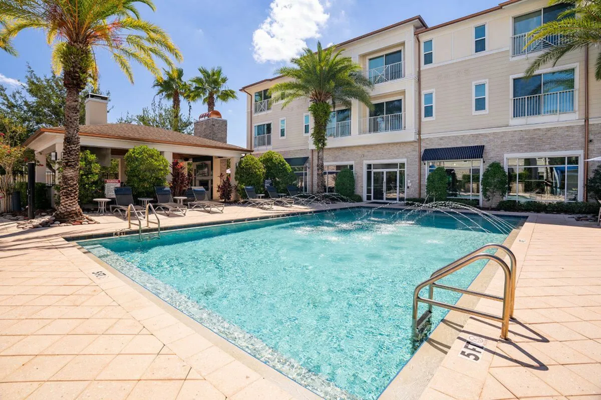 Luxurious apartment complex featuring a stunning swimming pool with elegant lounge chairs, surrounded by lush palm trees and modern architecture at The Marq Highland Park Apartments in Westchase Tampa