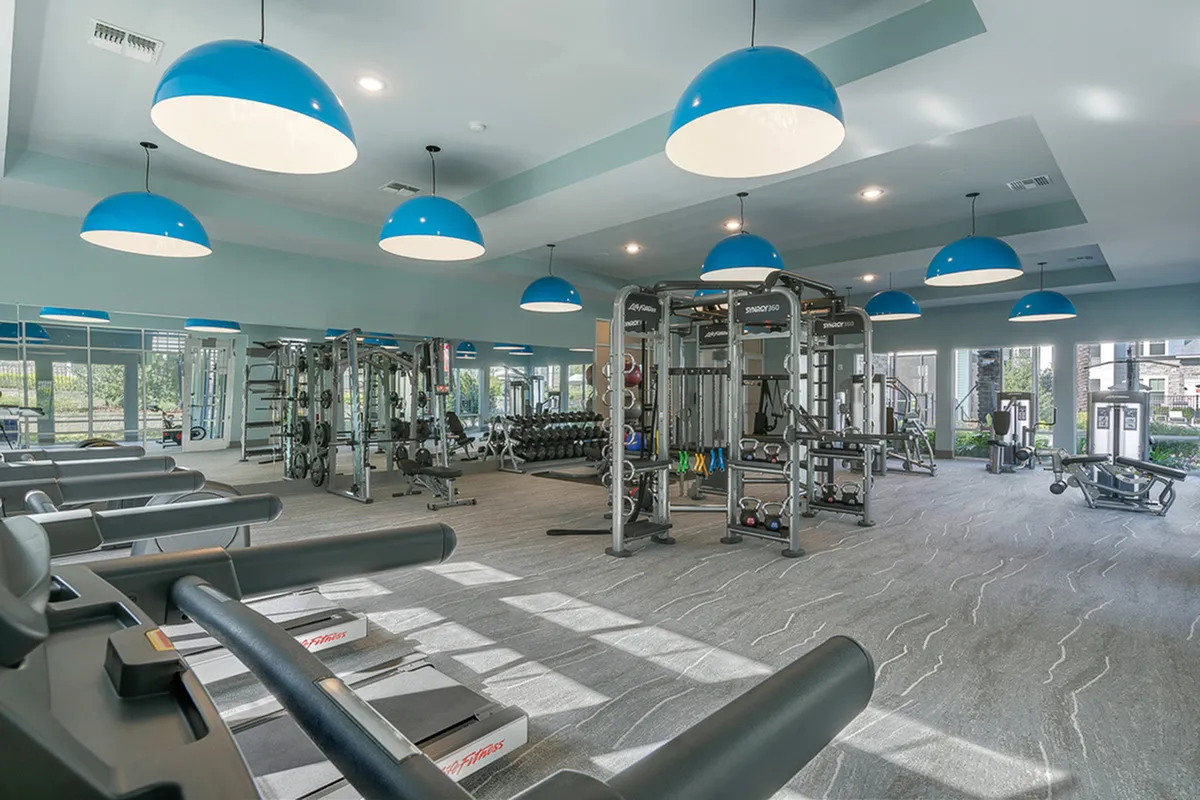 Spacious and modern fitness center at Cottonwood Clermont with state-of-the-art equipment, vibrant blue pendant lighting, and large windows for natural light.
