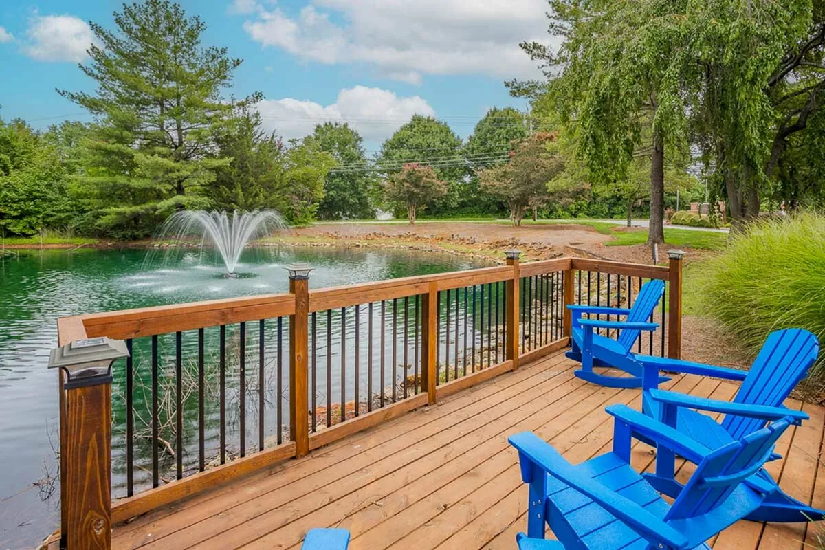 Charming wooden deck with vibrant blue chairs overlooking a serene pond with a graceful fountain and lush greenery.