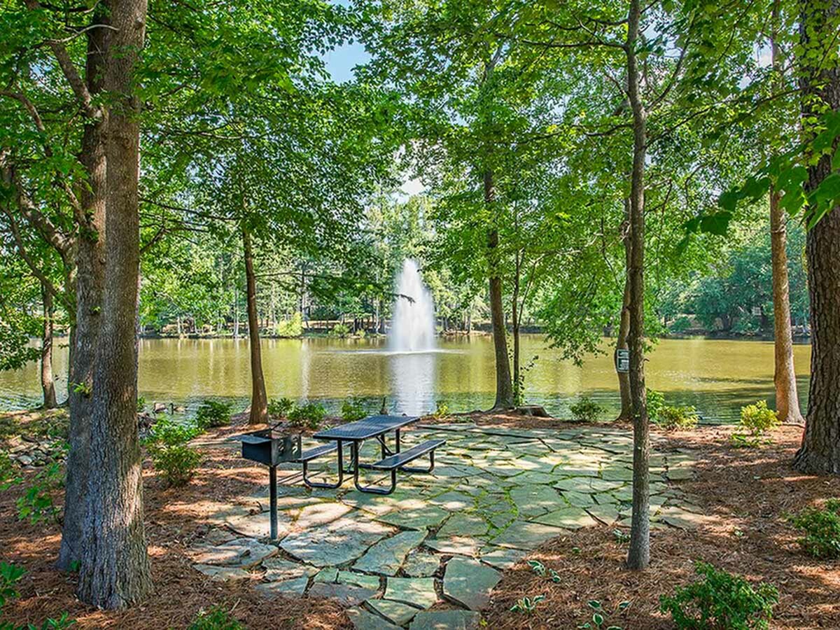 Serene outdoor setting with a shaded picnic area and tranquil lake view, featuring a picturesque fountain amidst lush greenery.