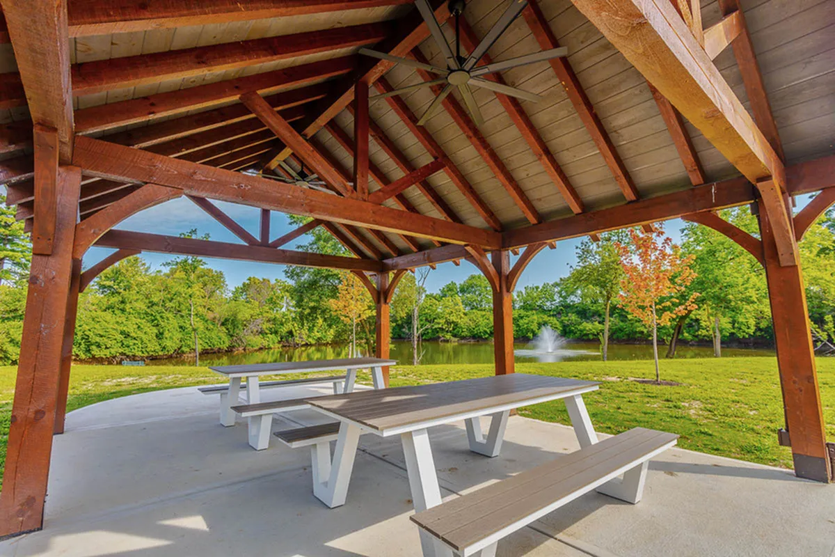 Charming outdoor pavilion with wooden beams, nestled by a serene pond and fountain, surrounded by lush greenery and vibrant trees.