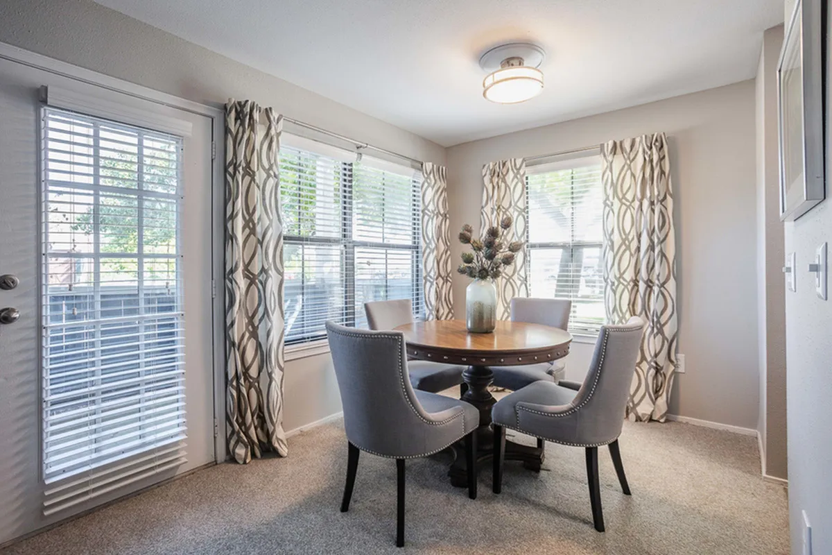 Bright dining room with elegant gray chairs surrounding a wooden table, enhanced by a modern overhead light and patterned curtains.