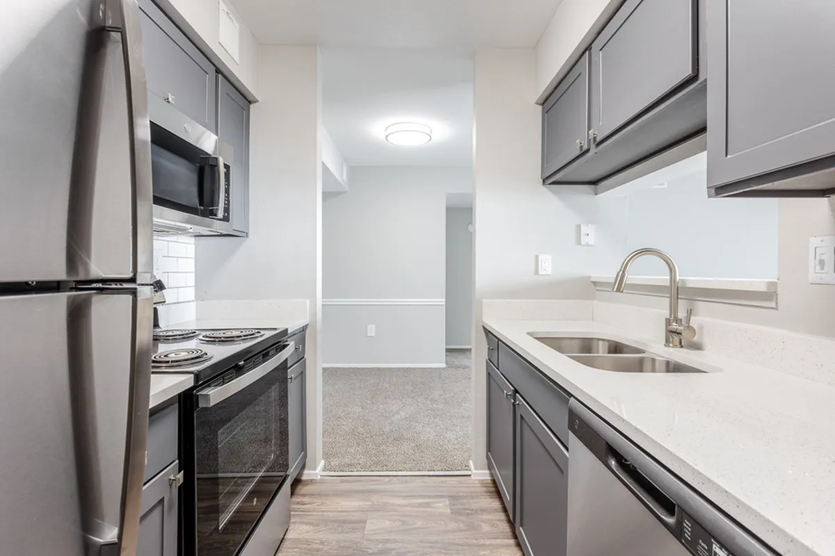 Modern apartment kitchen with sleek stainless steel appliances, elegant gray cabinetry, and pristine white countertops overlooking a cozy open-concept living space.