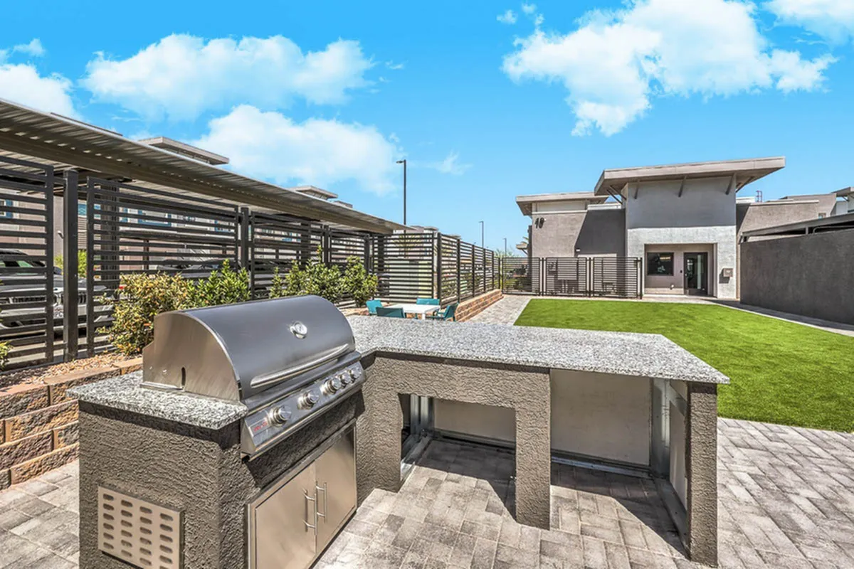 Modern outdoor kitchen with a built-in stainless steel grill, set in a stylish patio area with lush green lawn and contemporary building in the background under a bright blue sky.