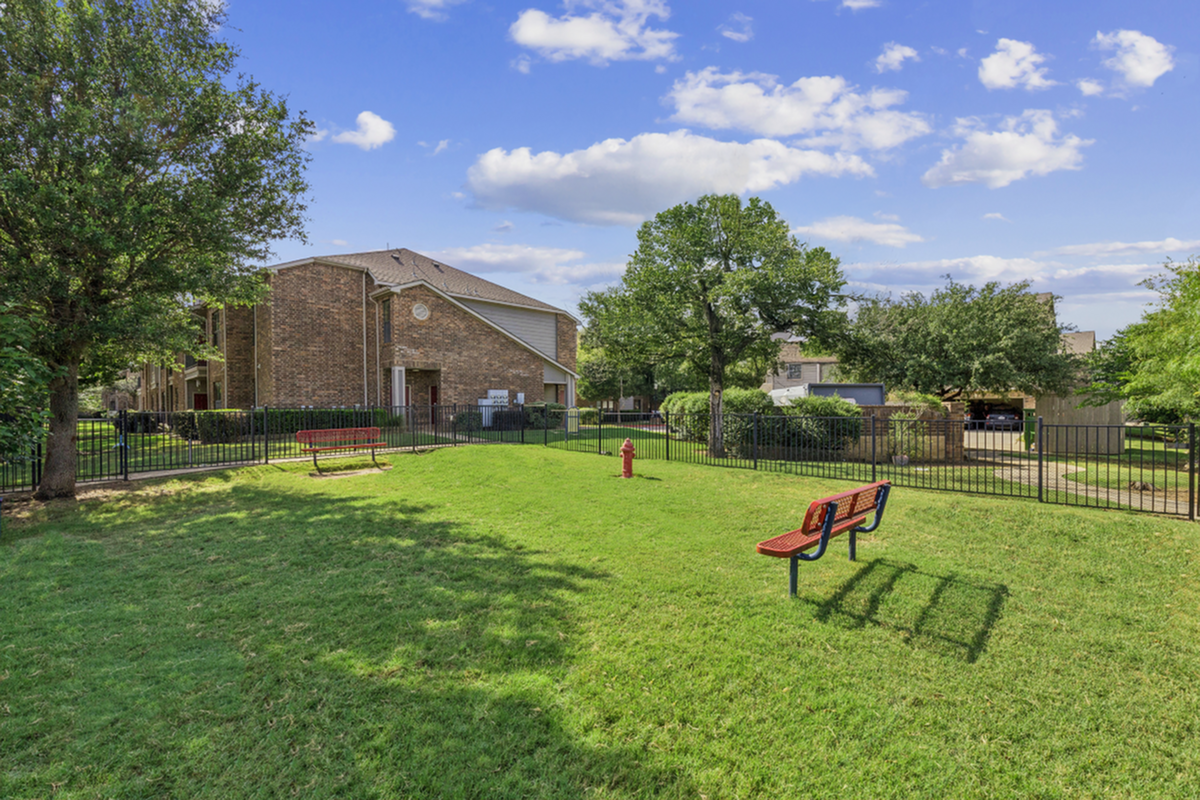 Charming apartment complex with lush green lawns, outdoor seating, and picturesque trees under a clear blue sky.