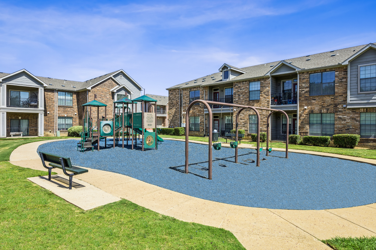 Charming apartment community with well-maintained playground, lush green spaces, and inviting brick exteriors under a bright blue sky.