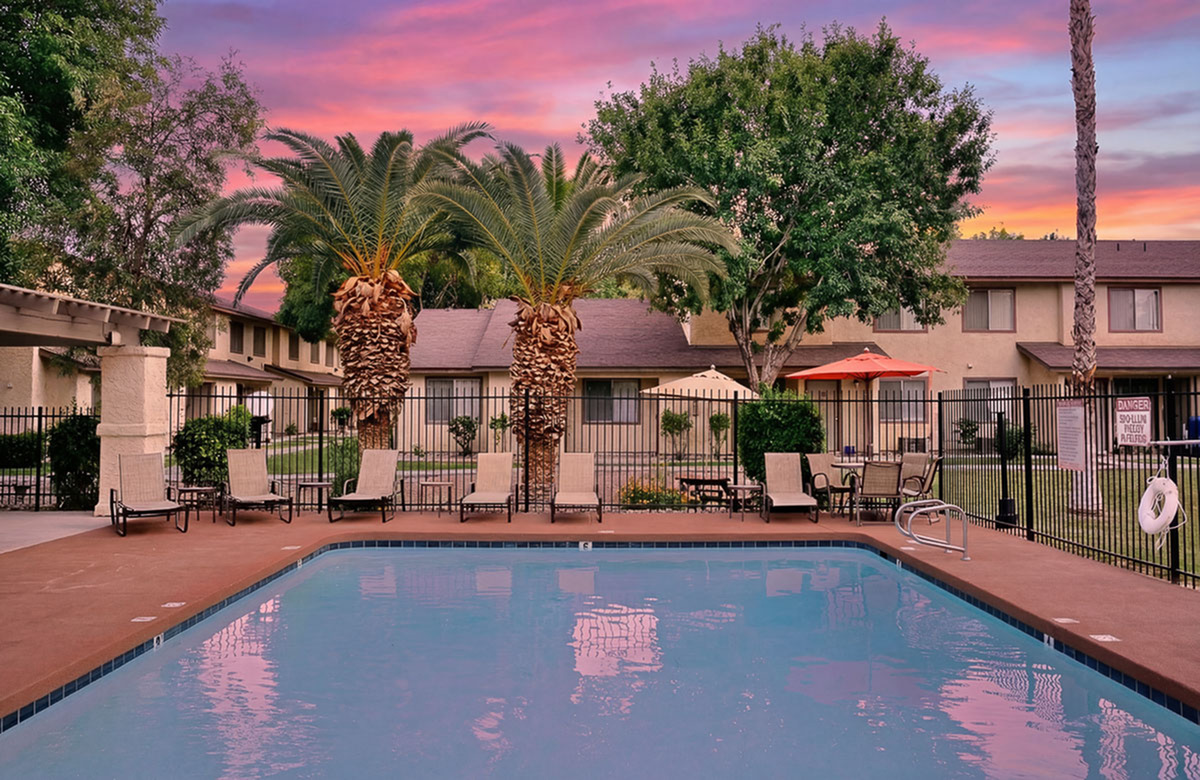 Charming apartment community with a refreshing pool, lush palm trees, and inviting lounge chairs under a vibrant sunset sky.