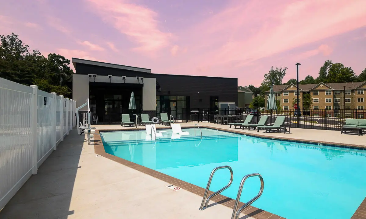 Luxurious outdoor pool area with modern lounge chairs, surrounded by a sleek fence and elegant building, set against a beautiful pink sunset sky.