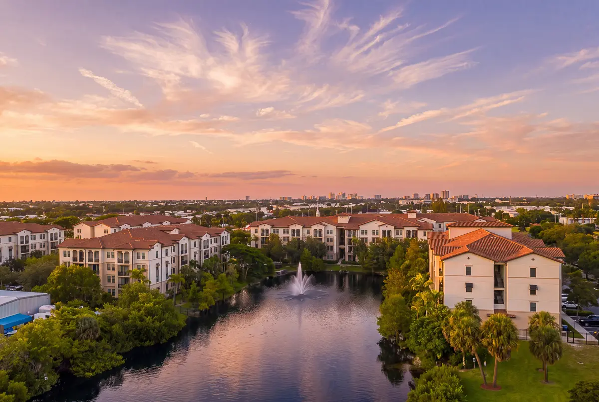 Scenic apartment complex with elegant architecture, surrounded by lush greenery and a tranquil lake at sunset.