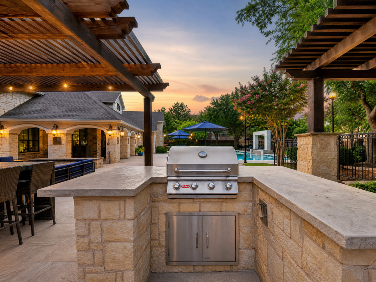 Luxurious outdoor patio featuring a modern grill, elegant stone countertops, and a stunning view of a lush garden and inviting pool at sunset at Toscana at Valley Ridge Apartments in Lewisville.