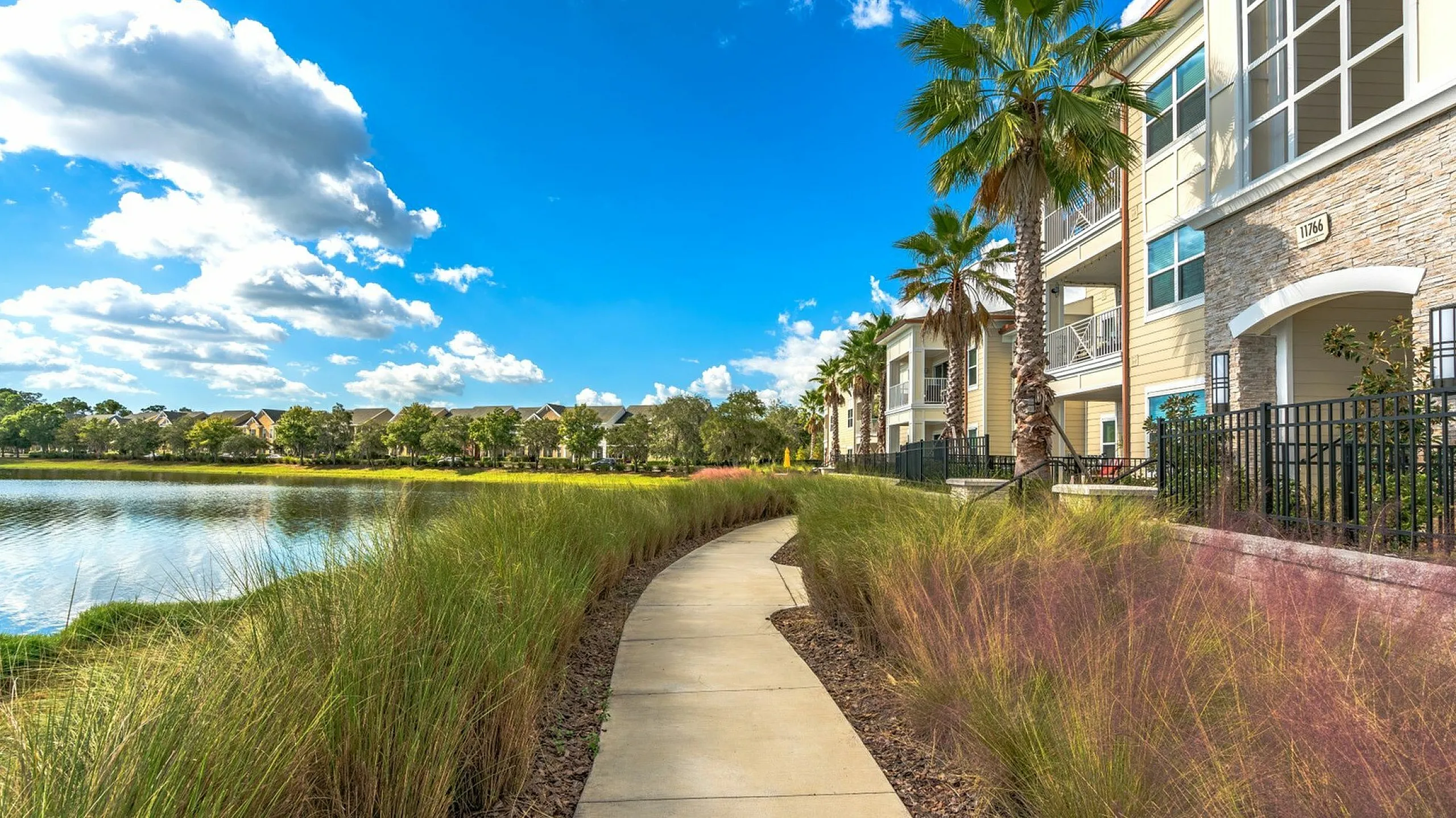 Scenic pathway alongside picturesque lake and luxury apartment buildings with palm trees.