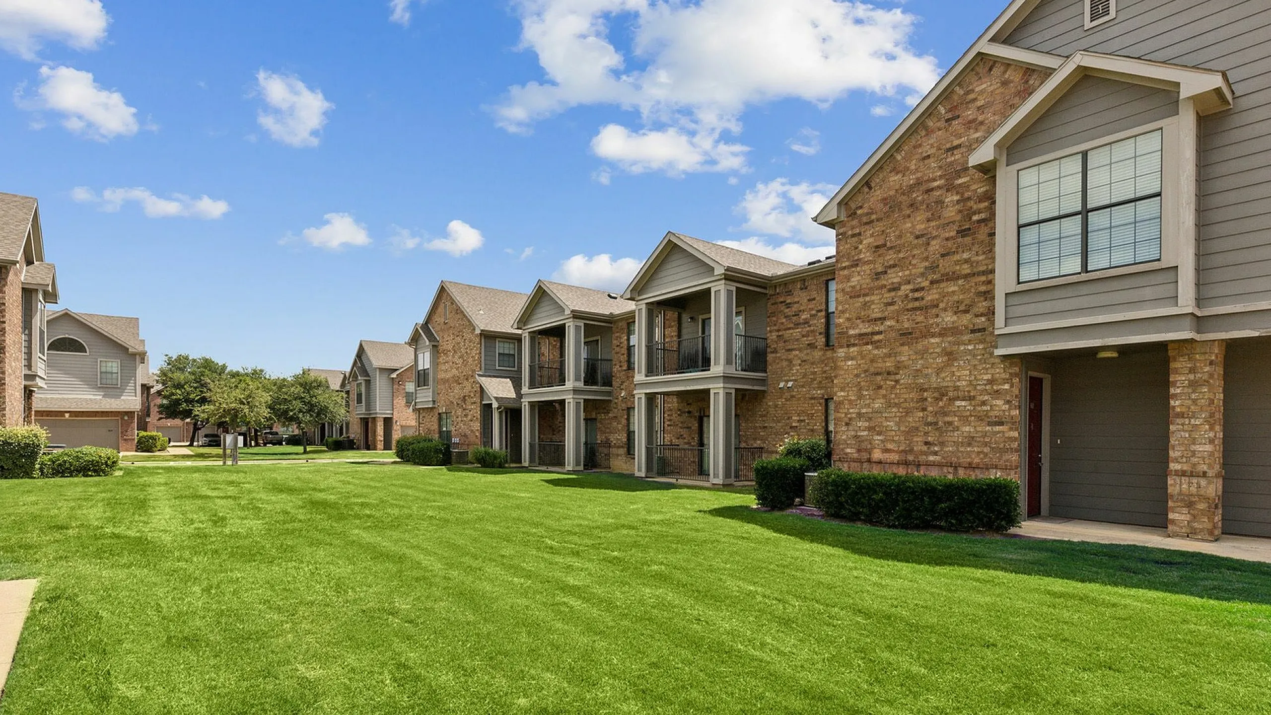 Charming brick apartment complex with spacious balconies, lush green lawns, and serene suburban setting under a clear blue sky.